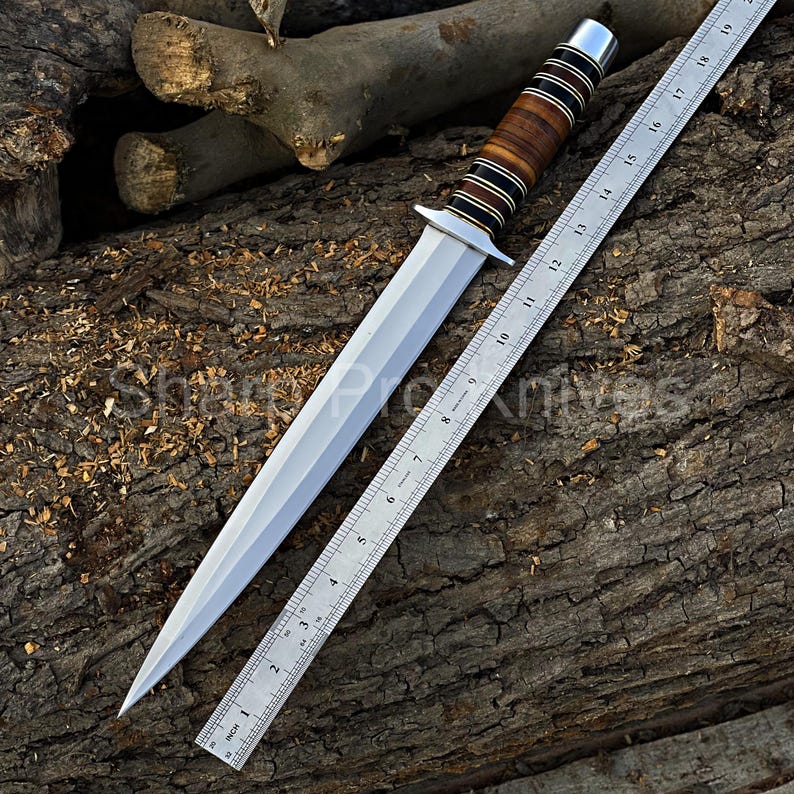 May include: A dagger with a polished steel blade and a handle featuring alternating bands of brown wood and black material. A metal ruler, marked in inches, is positioned alongside the blade. The dagger's total length is approximately 12 inches.
