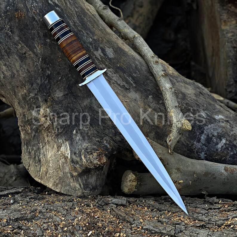 May include: A dagger with a polished silver blade and a handle featuring alternating bands of brown leather and black and silver metal. The text "Sharp Pro Knives" is visible on the wood background.