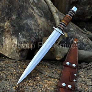 May include: A silver dagger with a double-edged blade and a brown and black leather-wrapped handle. The dagger is displayed with a brown leather sheath. The text "Shop Pro Knives" is visible.