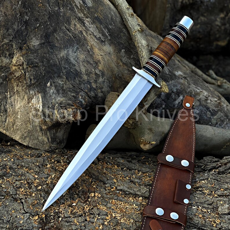 May include: A silver dagger with a double-edged blade and a brown and black leather-wrapped handle. The dagger is displayed with a brown leather sheath. The text "Shop Pro Knives" is visible.