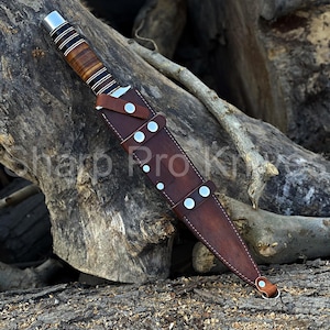 May include: A hunting knife with a brown wooden handle and a leather sheath. The knife has a silver bolster and a blade. The sheath is secured with silver rivets. The text "Sharp Pro Knives" is visible.