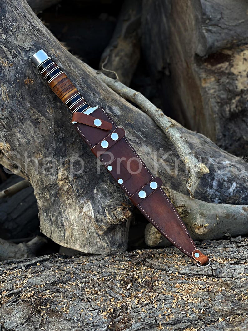 May include: A hunting knife with a brown wooden handle and a leather sheath. The knife has a silver bolster and a blade. The sheath is secured with silver rivets. The text "Sharp Pro Knives" is visible.
