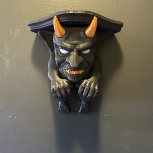 May include: A black gargoyle shelf with orange horns, white eyes, and orange teeth. The shelf is mounted on a gray wall. The gargoyle has its hands on its knees and is looking forward.