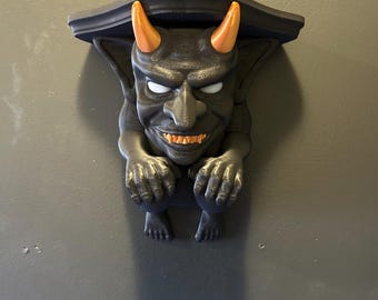 May include: A black gargoyle shelf with orange horns, white eyes, and orange teeth. The shelf is mounted on a gray wall. The gargoyle has its hands on its knees and is looking forward.