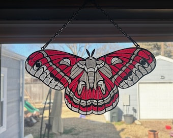 Puede incluir: Un atrapasoles de mariposa de vidriera con un diseño de alas rojas y blancas, colgado de una cadena. La mariposa tiene un patrón detallado y se muestra frente a una ventana, mostrando la vista exterior.