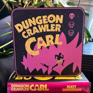 May include: A black and pink square coaster with the text "DUNGEON CRAWLER CARL" in yellow. The coaster features a running figure and a dog silhouette. Three skull icons are on the right side. The coaster is on top of a book with the same title.
