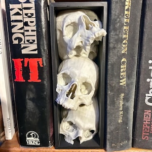 May include: A black rectangular display case holds three white skull sculptures. The skulls are stacked vertically, with visible eye sockets and nasal cavities. The display case is positioned between two Stephen King books on a wooden shelf.