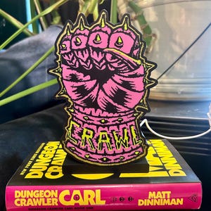 May include: A pink and black fist-shaped sticker with yellow accents and the word "CRAWL" in yellow. The sticker is on a black base, resting on a book titled "Dungeon Crawler Carl" by Matt Dinniman.