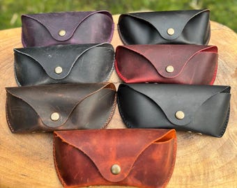 May include: A collection of handcrafted leather eyeglass cases in various colors, including black, brown, burgundy, and purple. Each case features a snap closure and a curved design, showcasing the natural texture of the leather.