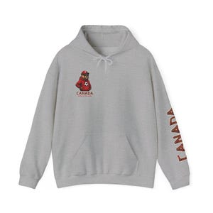 May include: A light gray hoodie with a kangaroo pocket and a drawstring hood. The front features a cartoon bear in a red jacket with the text "CANADA WORLD CUP 2026". The right sleeve has the word "CANADA" printed vertically in red.