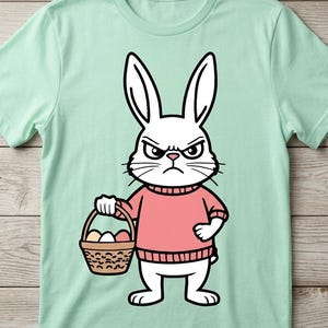 以下が含まれることがあります： ピンクのセーターを着てイースターエッグのバスケットを持っている、怒った白いウサギの漫画イラストが描かれた、明るい緑色のTシャツ。ウサギは厳しい表情をしており、黒で縁取られています。