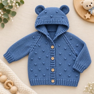 May include: A blue knitted cardigan featuring a hood with two small rounded ears on top. The garment is decorated with a textured bobble pattern across the front and sleeves, and it fastens down the centre with four light brown wooden buttons.