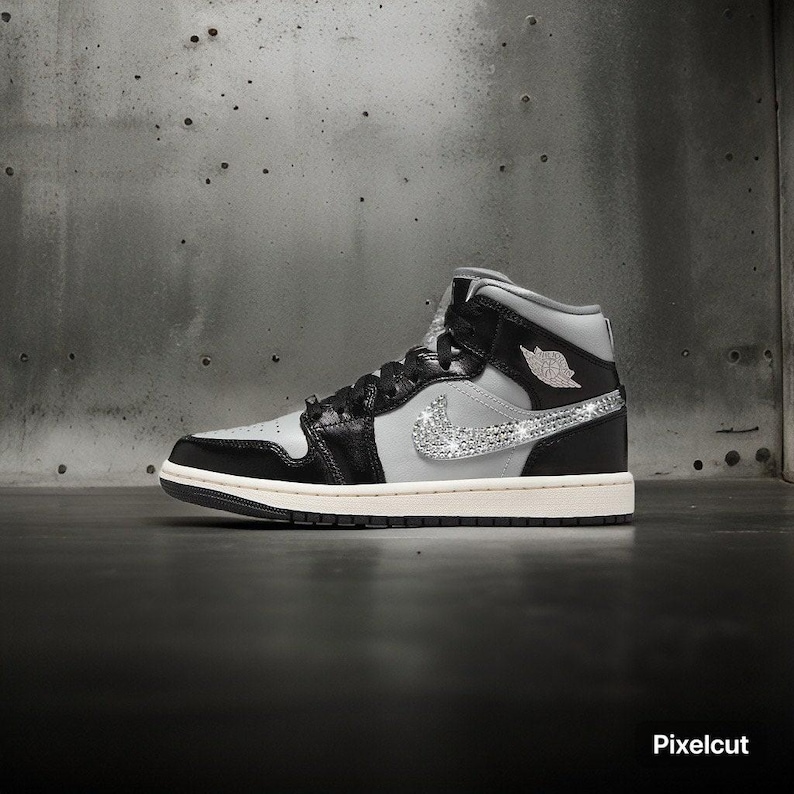 Op de afbeelding: Een high-top sneaker in zwart, grijs en wit met een sprankelende zilveren Nike swoosh. De schoen heeft zwarte veters, een witte zool en het Air Jordan-logo aan de zijkant. De sneaker staat tegen een grijze achtergrond.