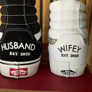 May include: A pair of black and white high-top sneakers with the words "Husband Est 2023" and "Wife Est 2023" written on the back of each shoe.