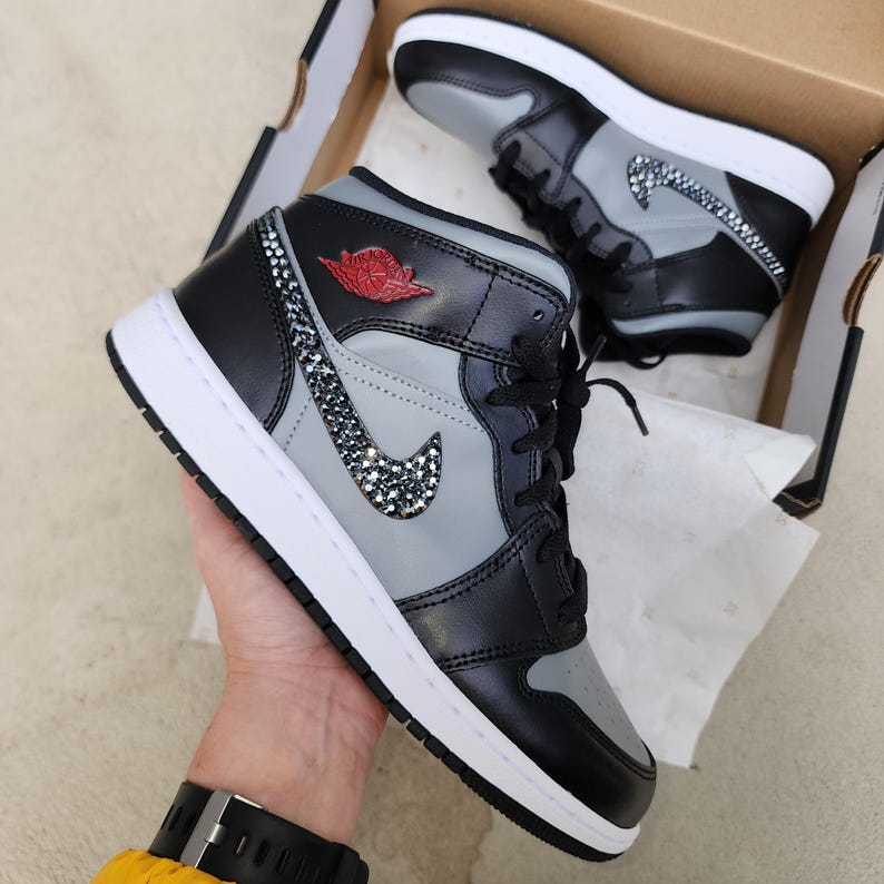 Op de afbeelding: Zwarte, grijze en witte high-top sneakers met kristallen versieringen. De sneakers hebben een grijs middenpaneel, zwarte overlays en een witte zool. Een rood logo is zichtbaar. De schoenen zitten in een kartonnen doos.