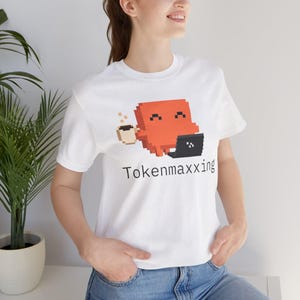 Może przedstawiać: Biały t-shirt z krótkim rękawem z grafiką pixel art przedstawiającą pomarańczową postać trzymającą kubek kawy i korzystającą z czarnego laptopa. Poniżej grafiki znajduje się napis Tokenmaxxing wydrukowany prostą czarną czcionką.