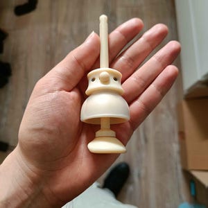 May include: A cream-colored, multi-part plastic component featuring a central vertical rod, a rounded mid-section with two small metallic screws on opposite sides, and a flared base. The object has a smooth, matte finish and a symmetrical design.