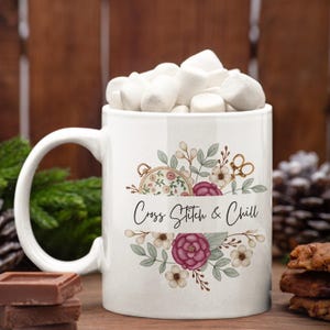 May include: White ceramic mug filled with marshmallows, featuring a floral design with a cross-stitch theme. The mug has the text "Cross Stitch & Chill" and is surrounded by chocolate and pine cones.
