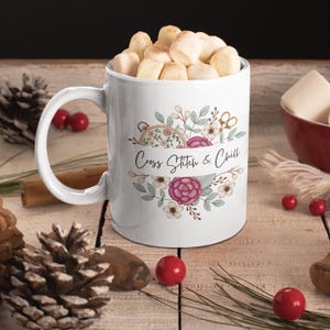 May include: White ceramic mug filled with marshmallows, featuring a floral design with the text "Cross Stitch & Chill." The mug has a large handle and is decorated with pink and white flowers, green leaves, and a cross-stitch hoop.