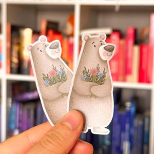Könnte beinhalten: Zwei Aufkleber mit Cartoon-Bären, die Blumensträuße halten. Die Bären sind hellbraun mit weißen Akzenten und einem lächelnden Gesicht. Die Blumen sind rosa, blau und grün. Die Aufkleber werden vor einem Bücherregal gehalten.