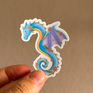Könnte beinhalten: Ein skurriler Aufkleber mit einem blauen Drachen mit gelbem Hals und lila Flügeln. Der Drache ist mit kleinen rosa Blumen geschmückt und hat ein lächelndes Gesicht. Der Aufkleber wird zwischen Fingern vor einem neutralen Hintergrund gehalten.
