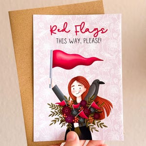 Könnte beinhalten: Eine Grußkarte mit hellrosa Hintergrund und weißem Blumenmuster. Der Text lautet Red Flags THIS WAY, PLEASE! Eine Illustration zeigt eine Person mit roten Haaren, die eine rote Flagge und einen Strauß dunkler Blumen mit einem schwarzen Vogel hält.