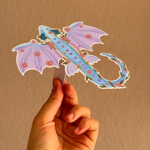 Könnte beinhalten: Ein dekorativer Drachenaufkleber mit hellblauem Körper, lavendelfarbenen Flügeln und einem geschwungenen Schwanz. Der Drache ist mit rosa Blüten und grünen Blättern verziert. Der Aufkleber wird vor einem neutralen Hintergrund gehalten.