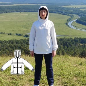 May include: A person wearing a white hooded jacket and navy blue pants stands on a grassy hill. The jacket has a zipper closure and pockets. The image includes a diagram of the jacket and text that reads "Sportswear sizes from US 6 / ES 38 to US 16 / ES 48."
