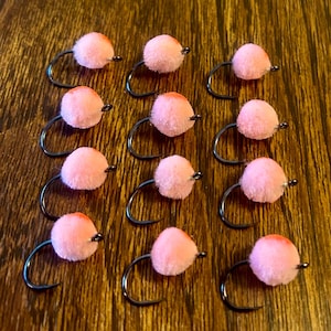 May include: Close-up of a collection of fishing lures. Each lure features a pink, fuzzy ball with an orange top, attached to a curved, black hook. The lures are arranged on a wooden surface, showcasing their design and color.