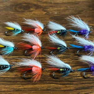 May include: A collection of ten fishing flies with various color combinations. Each fly features a black hook, white and gray feathering, and accents of yellow, red, orange, blue, and purple. The flies are arranged on a wooden surface.
