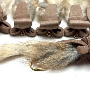 May include: A close-up of six brown foam fly fishing lures with white and brown fur tails. The lures are tied to gold hooks.