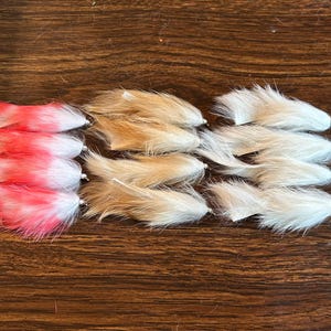 May include: A collection of fly fishing lures in three color variations: red and white, tan, and white. The lures are arranged in rows on a wooden surface, showcasing their fluffy, textured appearance.