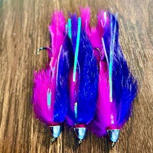 May include: Three fishing lures with vibrant pink and blue synthetic fibers. Each lure has a silver-colored head and a hook. Thin, light blue strips run down the center of each lure. The lures are arranged on a wooden surface.