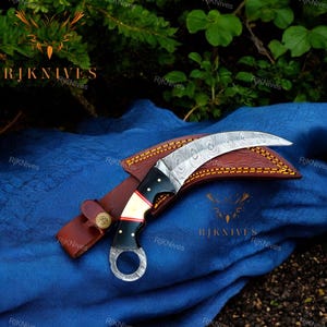 May include: A curved, Damascus steel karambit knife with a black handle and a red and white accent. The knife is in a brown leather sheath with a yellow stitching detail.