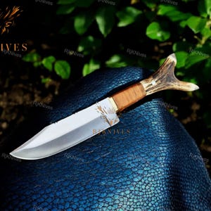 May include: A hunting knife with a brown wooden handle and a stag antler pommel. The blade is silver and has a slight curve. The knife is lying on a blue textured surface.