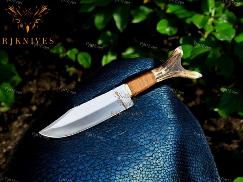May include: A hunting knife with a brown wooden handle and a stag antler pommel. The blade is silver and has a slight curve. The knife is lying on a blue textured surface.