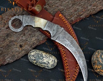 Può includere: Un coltello karambit in acciaio Damasco curvo con pomolo ad anello e manico in osso chiaro. Il coltello poggia su un fodero in pelle marrone con cuciture arancioni e un bottone circolare in ottone con un emblema a stella in rilievo.