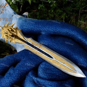 May include: A gold and silver sword with a detailed design and a sharp point. The sword is lying on a blue fabric background.