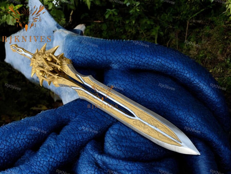 May include: A gold and silver sword with a detailed design and a sharp point. The sword is lying on a blue fabric background.