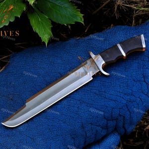 Könnte beinhalten: Ein großes, silberfarbenes Messer mit einem schwarzen Holzgriff. Die Klinge ist scharf und spitz, und der Griff hat einen strukturierten Griff. Das Messer liegt auf einer blauen Oberfläche.