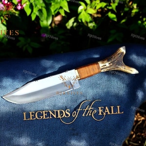 May include: A hunting knife with a brown wooden handle and a stag antler pommel. The blade is silver and has a slight curve. The knife is lying on a blue denim fabric with the words "Legends of the Fall" embroidered in gold.