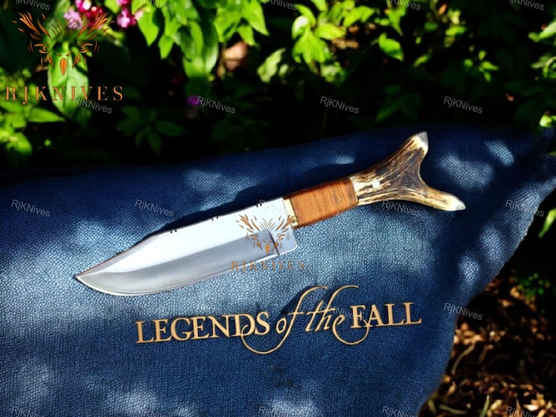 May include: A hunting knife with a brown wooden handle and a stag antler pommel. The blade is silver and has a slight curve. The knife is lying on a blue denim fabric with the words "Legends of the Fall" embroidered in gold.