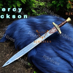 May include: A silver sword with a gold hilt and a brown handle. The sword is lying on a blue fuzzy surface. The text "Percy Jackson" is visible in the top left corner of the image.