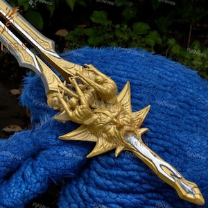 May include: A gold and silver sword with a detailed design of a winged creature with multiple figures. The sword is resting on a blue fuzzy surface.