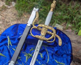 May include: A silver-toned rapier sword with a white handle and an ornate gold-colored hilt featuring small red accents. The sword is accompanied by a black scabbard with silver-colored metal fittings, both displayed on a vibrant blue fabric.