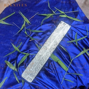 May include: A rectangular metal knife blade blank featuring a distinct feather-like Damascus steel pattern in black and white. The blade is positioned diagonally across a vibrant blue fabric surface decorated with scattered green leaves.