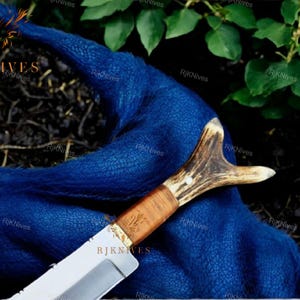 May include: A hunting knife with a brown antler handle and a silver blade. The knife is lying on a blue fabric background.
