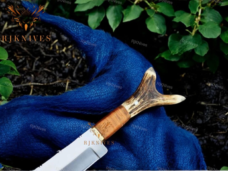 May include: A hunting knife with a brown antler handle and a silver blade. The knife is lying on a blue fabric background.