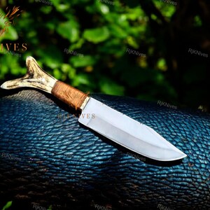May include: A hunting knife with a brown wooden handle and a white antler pommel. The blade is silver and has a slight curve. The knife is lying on a blue textured surface.