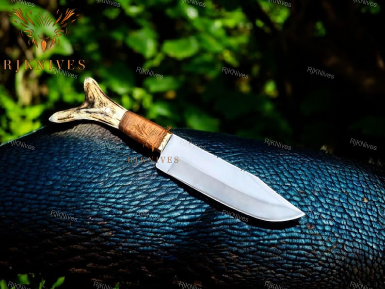 May include: A hunting knife with a brown wooden handle and a white antler pommel. The blade is silver and has a slight curve. The knife is lying on a blue textured surface.